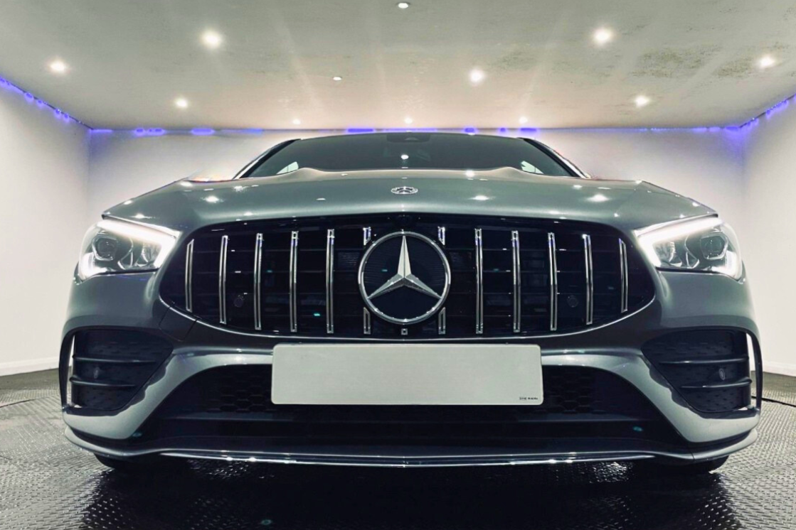 A silver car with a prominent grille and illuminated headlights is parked indoors under bright ceiling lights, creating a sleek and modern atmosphere. A Mercedes-Benz logo is centered on the grille.