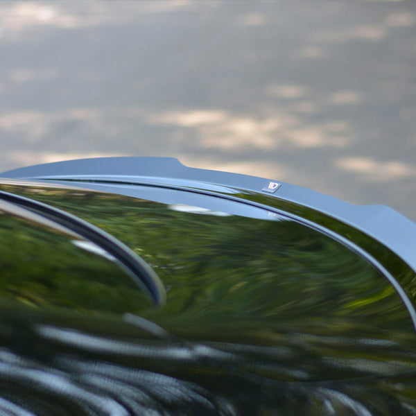 A car roof reflects blurred greenery and sky; its black surface is curved smoothly. In the background, the road appears indistinct through sunlight-dappled shadows.