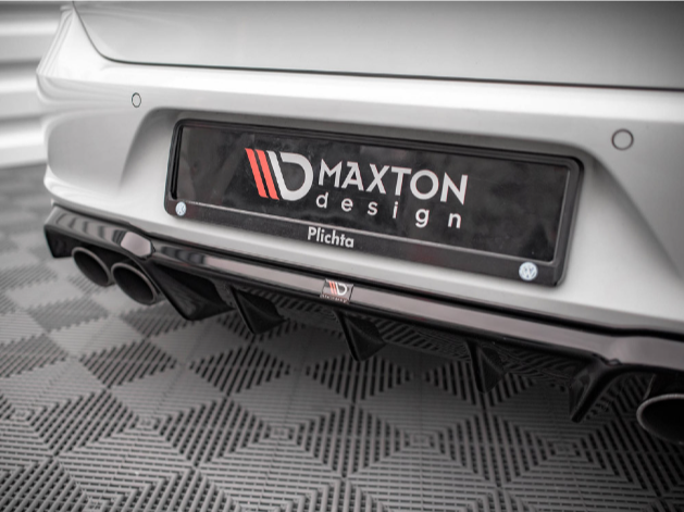 A car bumper features a license plate that reads "MAXTON design Plichta." Dual exhaust pipes are visible beneath. The car rests on a textured, patterned floor in a well-lit showroom.