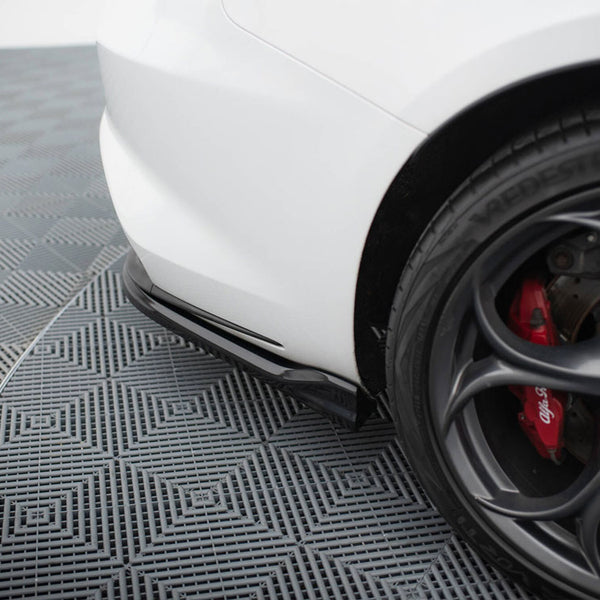 A white car's rear bumper is parked on a patterned grid floor. The black alloy wheel features red brake calipers labeled "Alfa Romeo." The tire brand reads "VREDESTEIN."