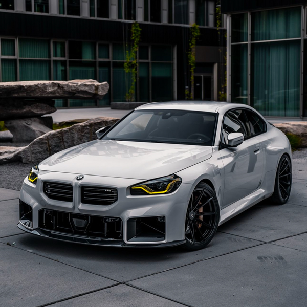 A sleek white sports car with yellow headlights is parked on a concrete surface, surrounded by modern glass buildings and large stone sculptures. BMW logo visible on the grille.