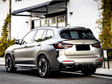 A metallic BMW iX3 parked on a street beside a building with glass doors and greenery. The rear of the car shows sleek taillights and 