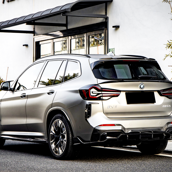 A metallic BMW iX3 parked on a street beside a building with glass doors and greenery. The rear of the car shows sleek taillights and "iX3" and other text on the back.