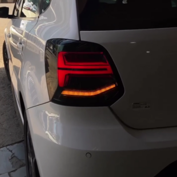 A white car's rear section with illuminated tail lights is parked near a building, casting reflections on its glossy surface.