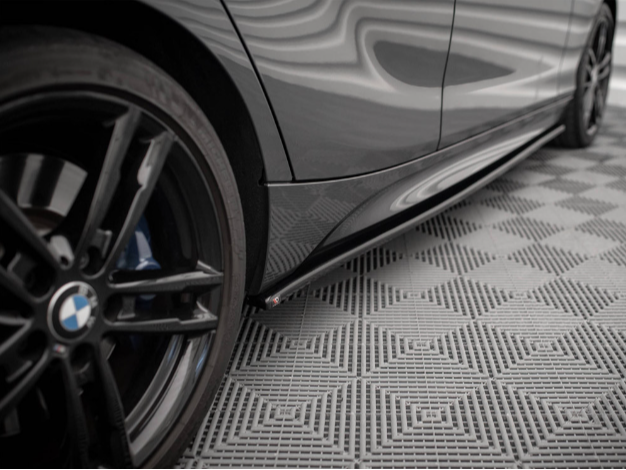 A black car tire is stationary next to a sleek vehicle body, reflecting a wavy pattern. The vehicle is parked on a textured, grid-like floor surface.