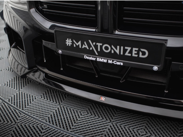 A car's front bumper is shown with a black license plate frame featuring the hashtag "#MAXTONIZED" above "Dealer BMW M-Cars." The car is parked on a patterned floor.