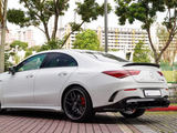 White Mercedes-Benz CLA 45 AMG parked on a patterned pavement, surrounded by trees and buildings in the background. The rear of the car features distinctive taillights and quad exhaust pipes.
