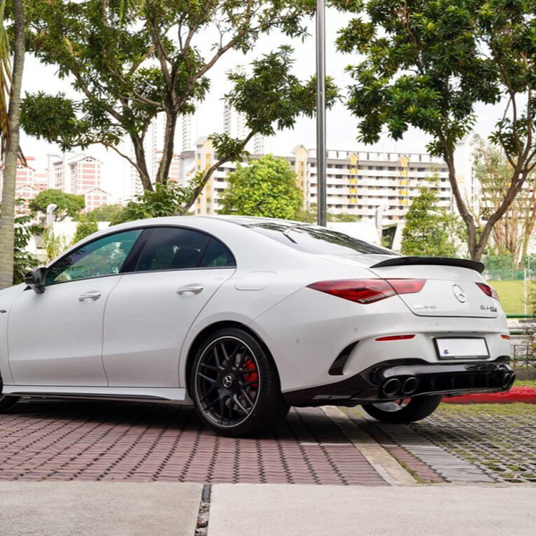 White Mercedes-Benz CLA 45 AMG parked on a patterned pavement, surrounded by trees and buildings in the background. The rear of the car features distinctive taillights and quad exhaust pipes.