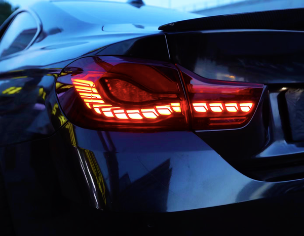 A car's taillight glows red in the evening light, with sleek body lines reflecting its urban surroundings. The context suggests an area with building reflections visible on the vehicle.