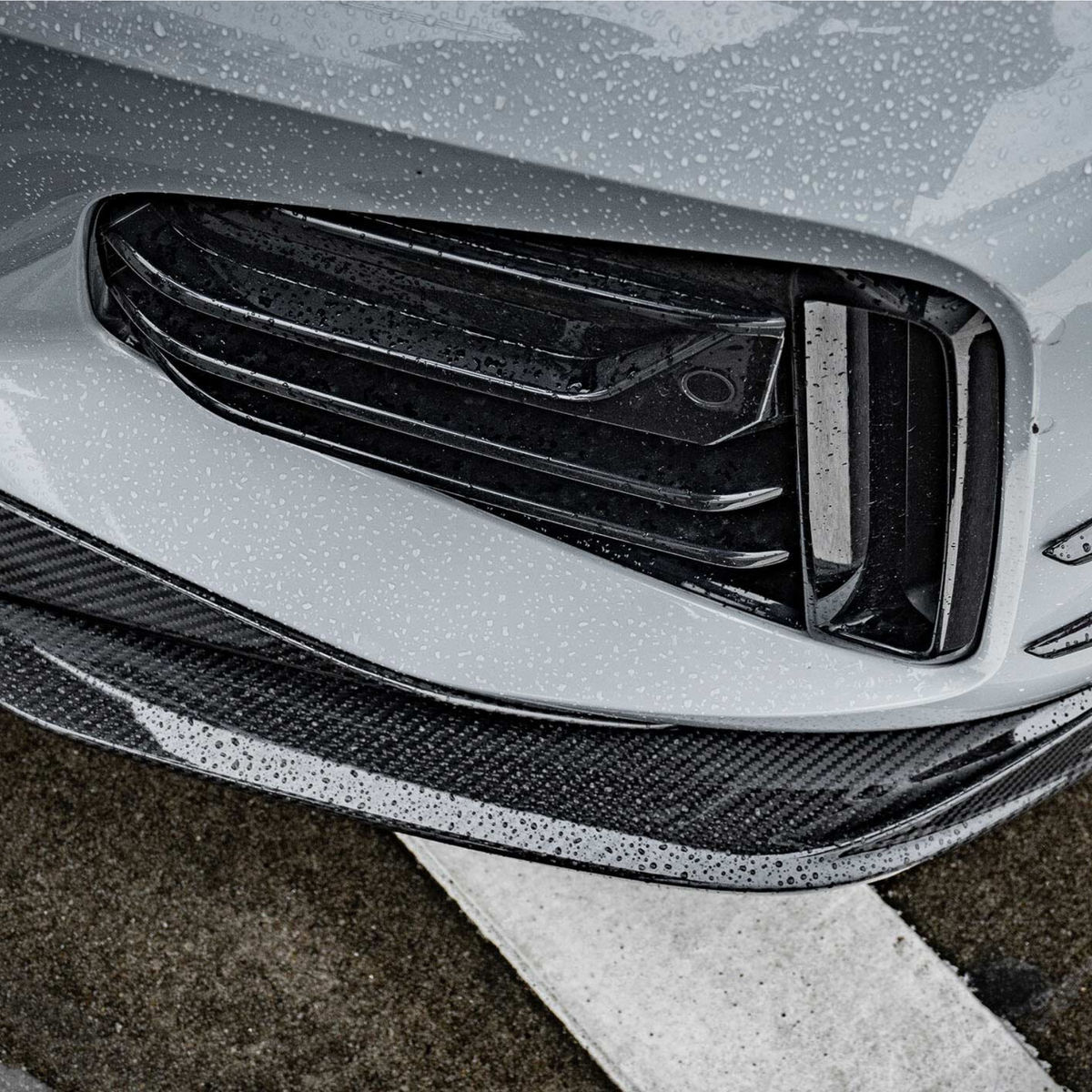 A car's front bumper, featuring sleek black grills and carbon fiber accents, is covered in rain droplets, situated on a wet asphalt surface with white road markings.