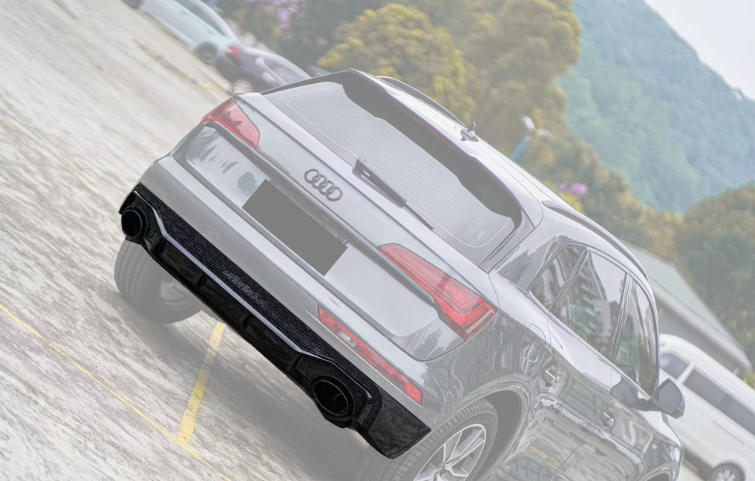 A gray Audi SUV is parked on a concrete lot, surrounded by other cars. Trees and hills create a natural backdrop. The rear badge features the Audi logo.