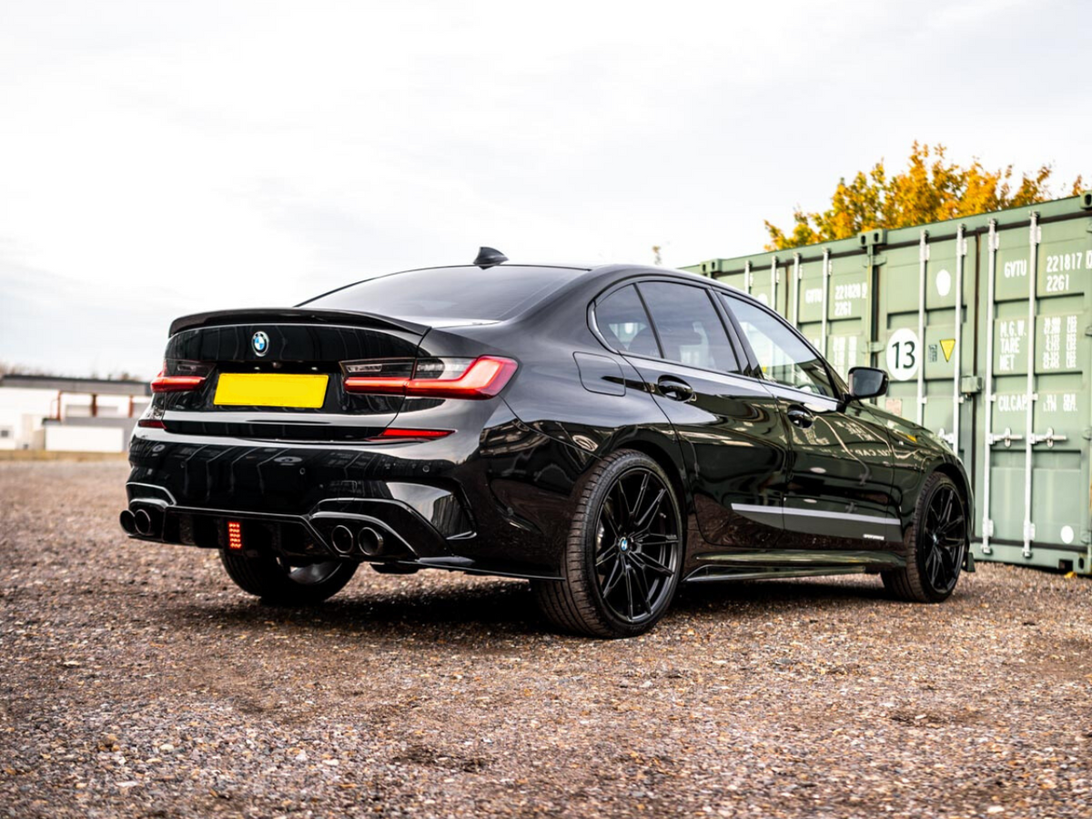 A sleek black car is parked on a gravel lot near green shipping containers. The car's rear features twin exhausts and shiny black rims, and the background has trees and buildings.