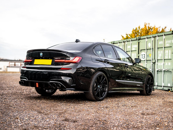 A sleek black car is parked on a gravel lot near green shipping containers. The car's rear features twin exhausts and shiny black rims, and the background has trees and buildings.