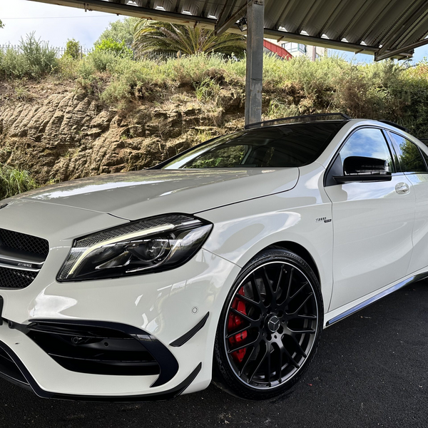 A white Mercedes-Benz car, parked under a metal shelter, features black alloy wheels and is set against a background of greenery and a rocky wall.