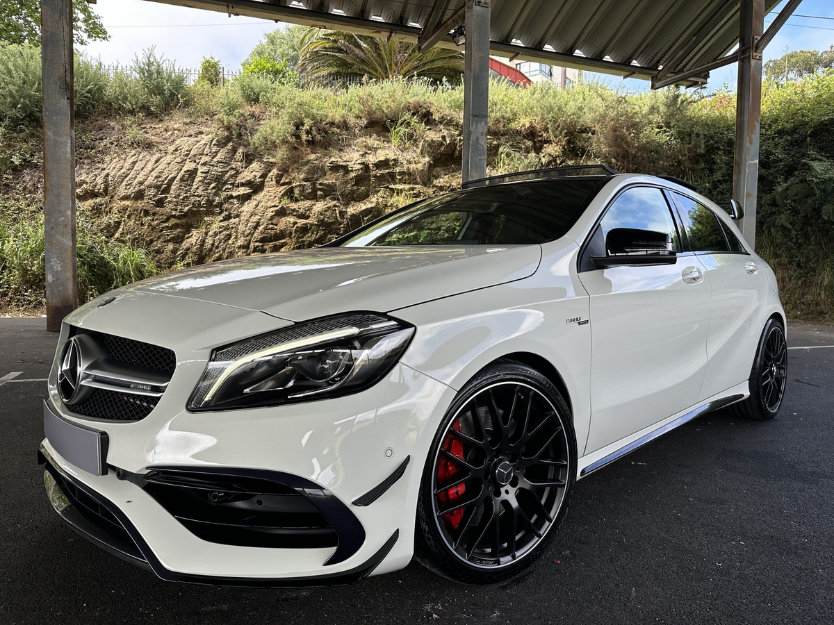 A white Mercedes-Benz car, parked under a metal shelter, features black alloy wheels and is set against a background of greenery and a rocky wall.