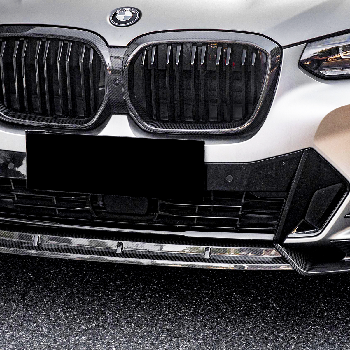 A BMW car front features a prominent grille and sleek headlights, parked on a textured asphalt surface. A black license plate obscures the lower section.