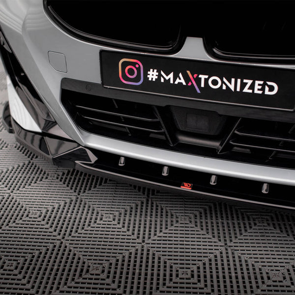 A car's front bumper with a black license plate holder, displaying "#MAXTONIZED" next to an Instagram logo, is parked on a textured gray floor.