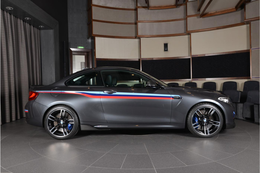 A gray sports car with red, blue, and white stripes is parked indoors on a tiled floor, surrounded by dark curtains and theater seating.