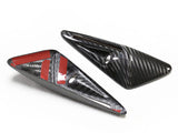 Two carbon fiber arrowhead-shaped objects, one featuring red adhesive strips, rest on a white surface. Each has a small circular hole, suggesting mounting capabilities.