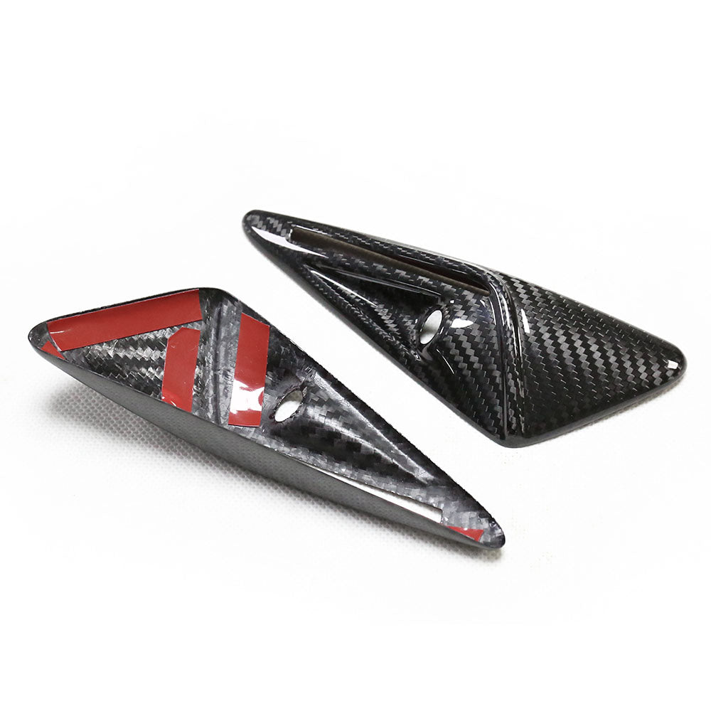 Two carbon fiber arrowhead-shaped objects, one featuring red adhesive strips, rest on a white surface. Each has a small circular hole, suggesting mounting capabilities.
