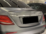 A silver Mercedes AMG E43 is parked indoors, showcasing badges on the trunk. The surrounding area has a modern, well-lit showroom with another vehicle visible nearby.