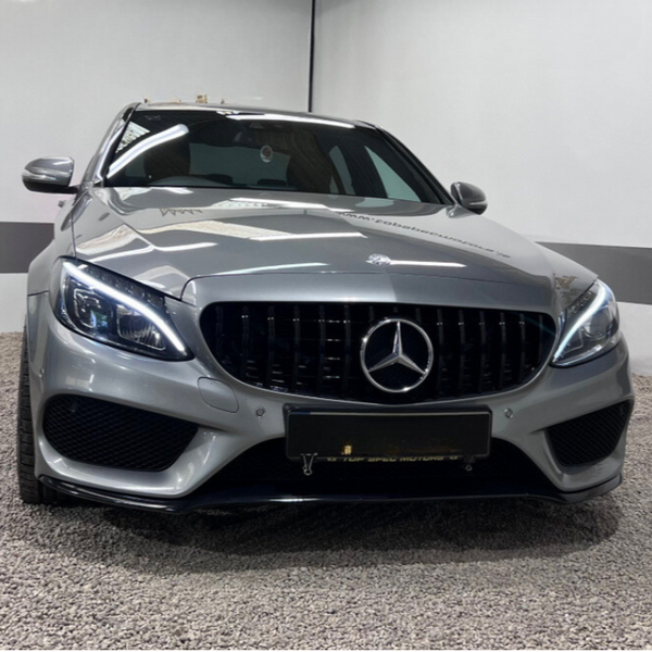 A silver car with illuminated headlights is parked indoors on a gravel floor, featuring a prominent emblem on the grille and reflective lighting in a simple white-walled setting.