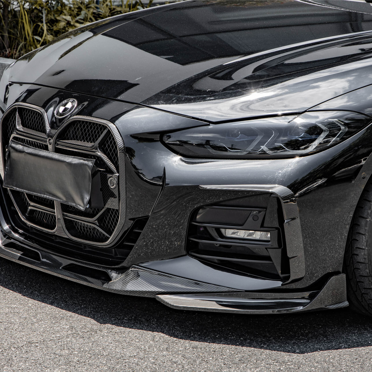 A sleek black car, parked on a street, with a distinctive front grille and stylish black wheels. It is positioned near a concrete pavement with greenery in the background.