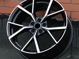 A black alloy wheel with a silver, angular spoke design leans against an orange brick wall on a dark pavement.