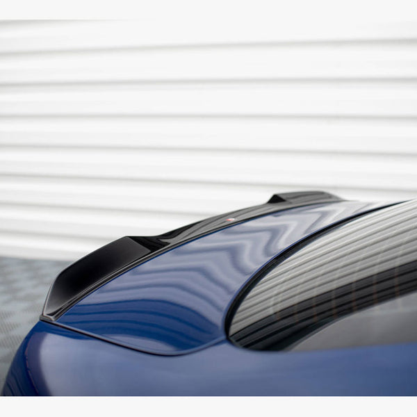 A sleek, blue car with a black spoiler rests on a textured surface, with a corrugated metal wall in the background reflecting on the rear window.