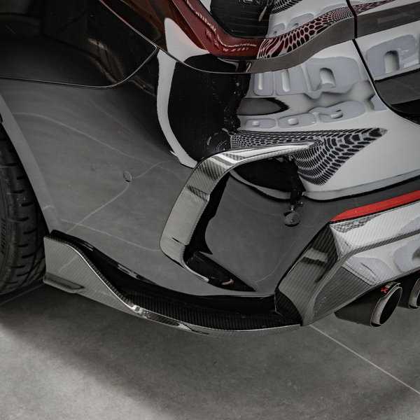 A sleek black car's rear corner, showcasing a tinted taillight and dual exhaust pipes, is parked indoors on a smooth gray floor.
