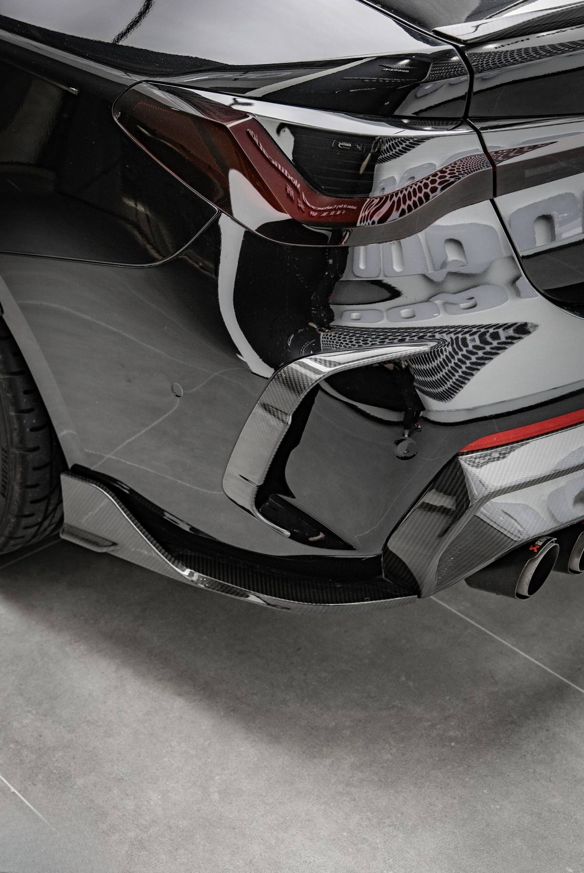 A sleek black car's rear corner, showcasing a tinted taillight and dual exhaust pipes, is parked indoors on a smooth gray floor.