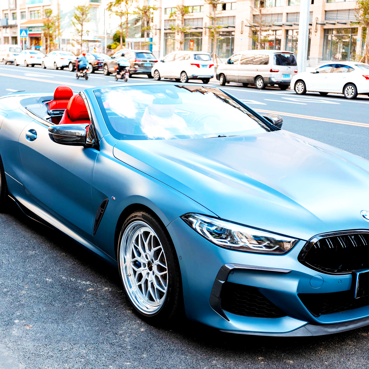A blue convertible car with red seats is parked on a city street, surrounded by traffic and buildings, under a clear sky.