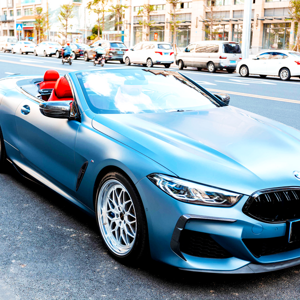 A blue convertible car with red seats is parked on a city street, surrounded by traffic and buildings, under a clear sky.