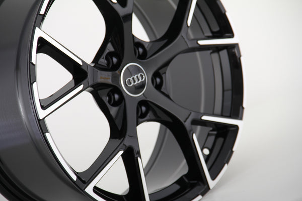 A black alloy car wheel rim, featuring five double spokes with a metallic silver outline, displays the Audi logo at the center. It is positioned against a plain white background.