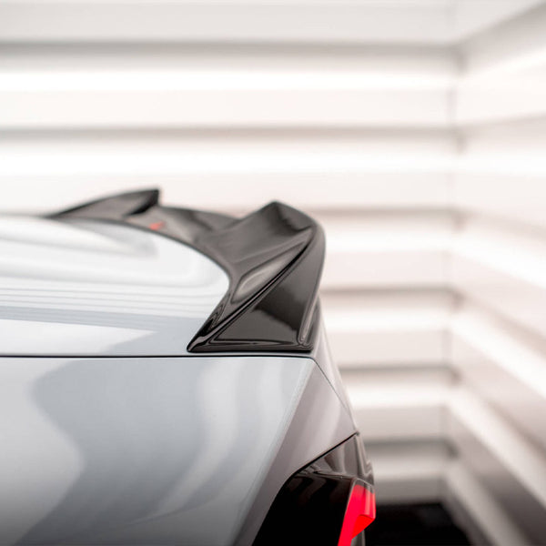 A car's rear spoiler is visible in close-up, featuring a glossy black finish. It is situated in a brightly lit, white-paneled room, enhancing its sleek design.