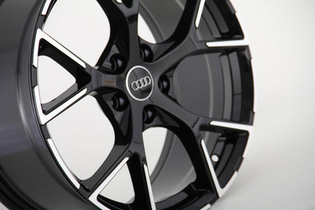 Black and silver alloy wheel, featuring a five-spoke design with the Audi logo at the center, set against a plain white background.