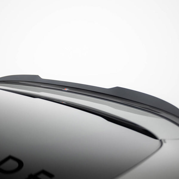 A black car roof spoiler is mounted on a smooth, grey vehicle surface. Partial letters "DE" are visible on the car, set against a plain, white background.