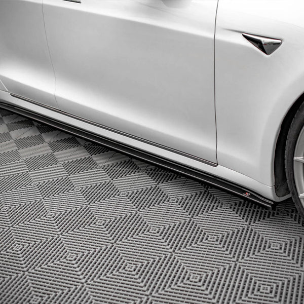 A white car with sleek, angular design rests stationary on a textured, diamond-patterned gray floor, showcasing its aerodynamic side skirts and alloy wheels in a well-lit indoor setting.