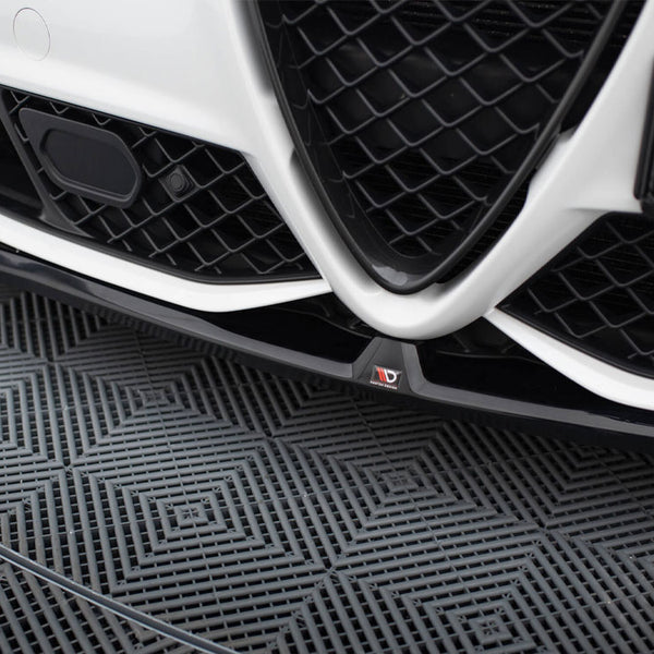 Car grille with a V-shaped design and black mesh pattern, positioned close to the ground. The left side shows a sensor. The floor has a textured geometric pattern.