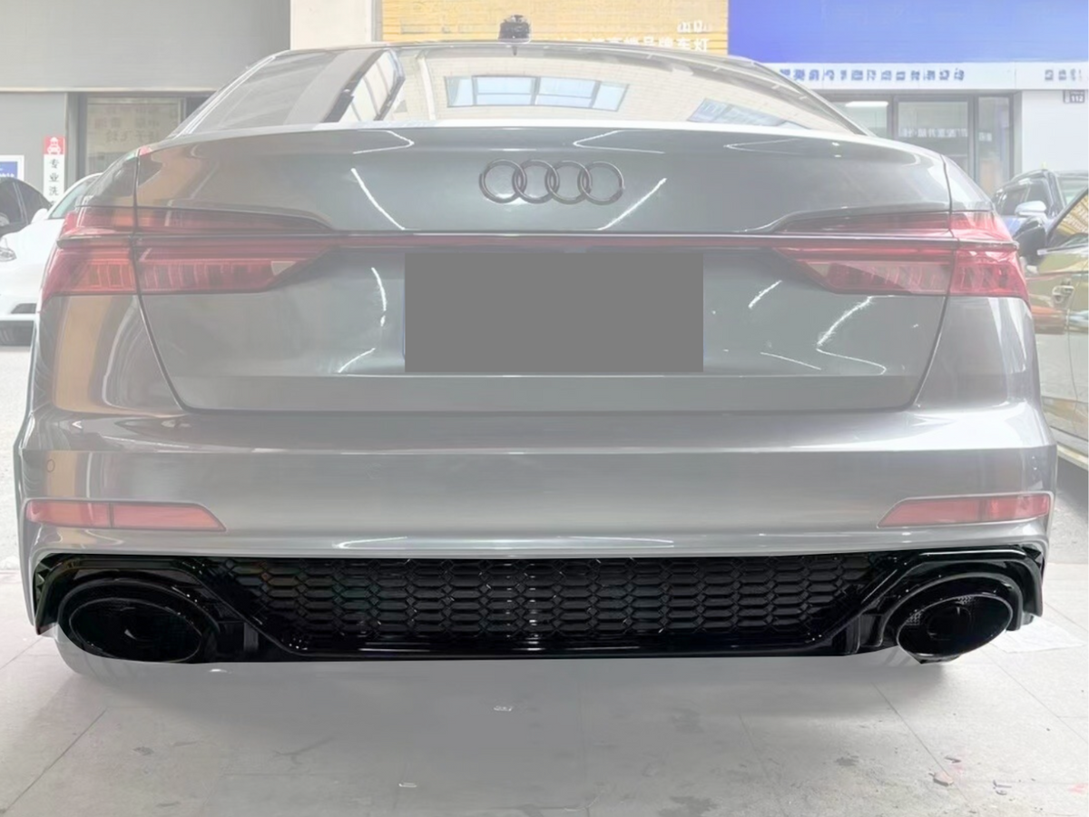 A silver Audi car is stationary inside a showroom, displaying its rear view with distinctive taillights and dual exhaust, parked among other cars under bright, reflective lighting.
