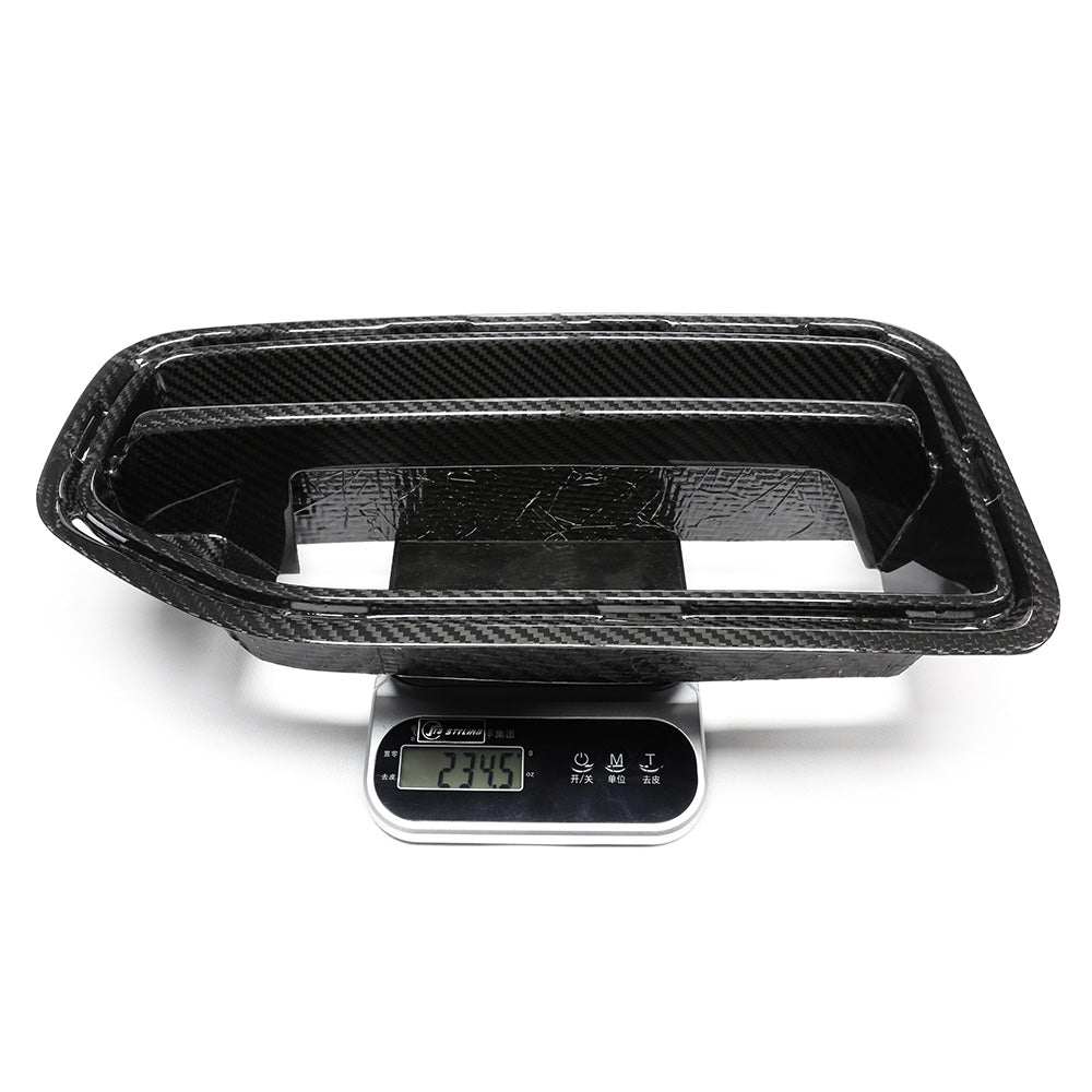 A black carbon fiber car part is placed on a digital scale displaying "234.5" in a well-lit, white studio setting.