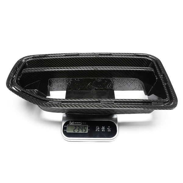 A black carbon fiber car part is placed on a digital scale displaying "234.5" in a well-lit, white studio setting.