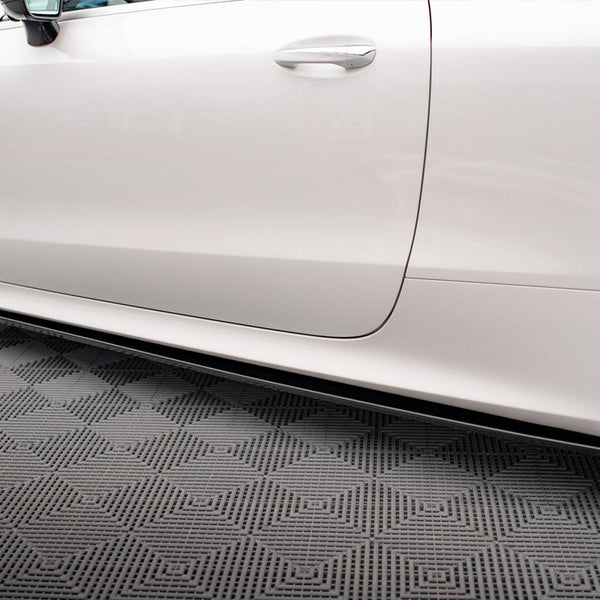 A white car is parked in a garage on a textured gray floor. The focus is on the side view, showing part of the front and rear tires.