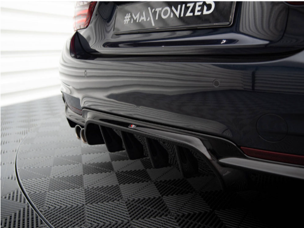 A car's rear bumper features a #MAXTONIZED license plate and quad exhaust pipes, set in a garage with a textured floor pattern and reflective panels.