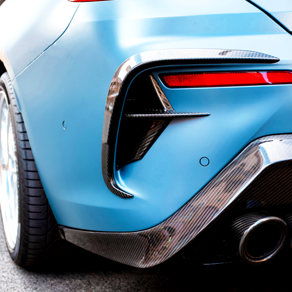 A blue sports car's rear features chrome exhausts and red tail lights, parked on a city street with another car partially visible in the background.
