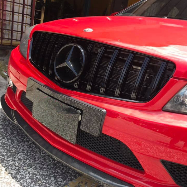 A red car features a prominent black grille with a Mercedes logo, parked on a sunlit, paved surface near a building with a metal door.