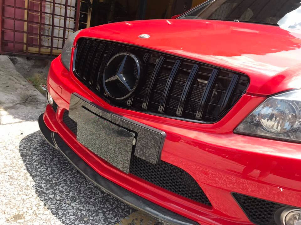 A red car features a prominent black grille with a Mercedes logo, parked on a sunlit, paved surface near a building with a metal door.