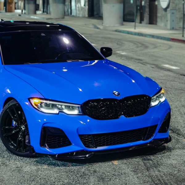 A blue BMW car is parked on a dimly lit street at night, with its headlights on, surrounded by urban infrastructure.