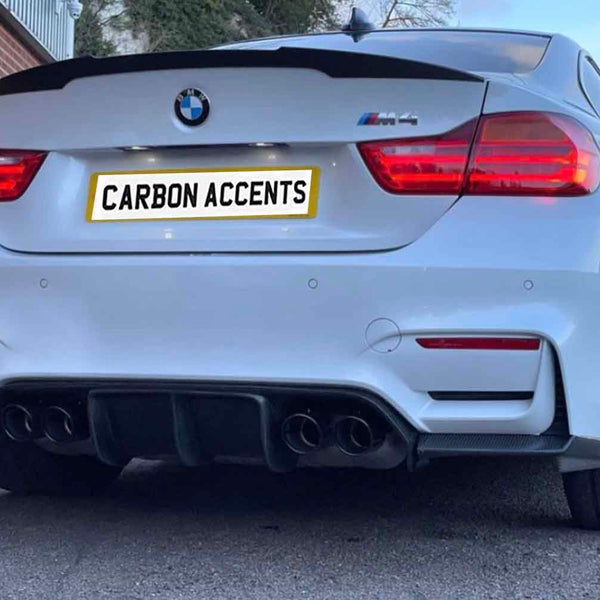 A white BMW M4 is parked, featuring dual exhausts and illuminated rear lights. The license plate reads "CARBON ACCENTS." Brick walls and a building are visible in the background.