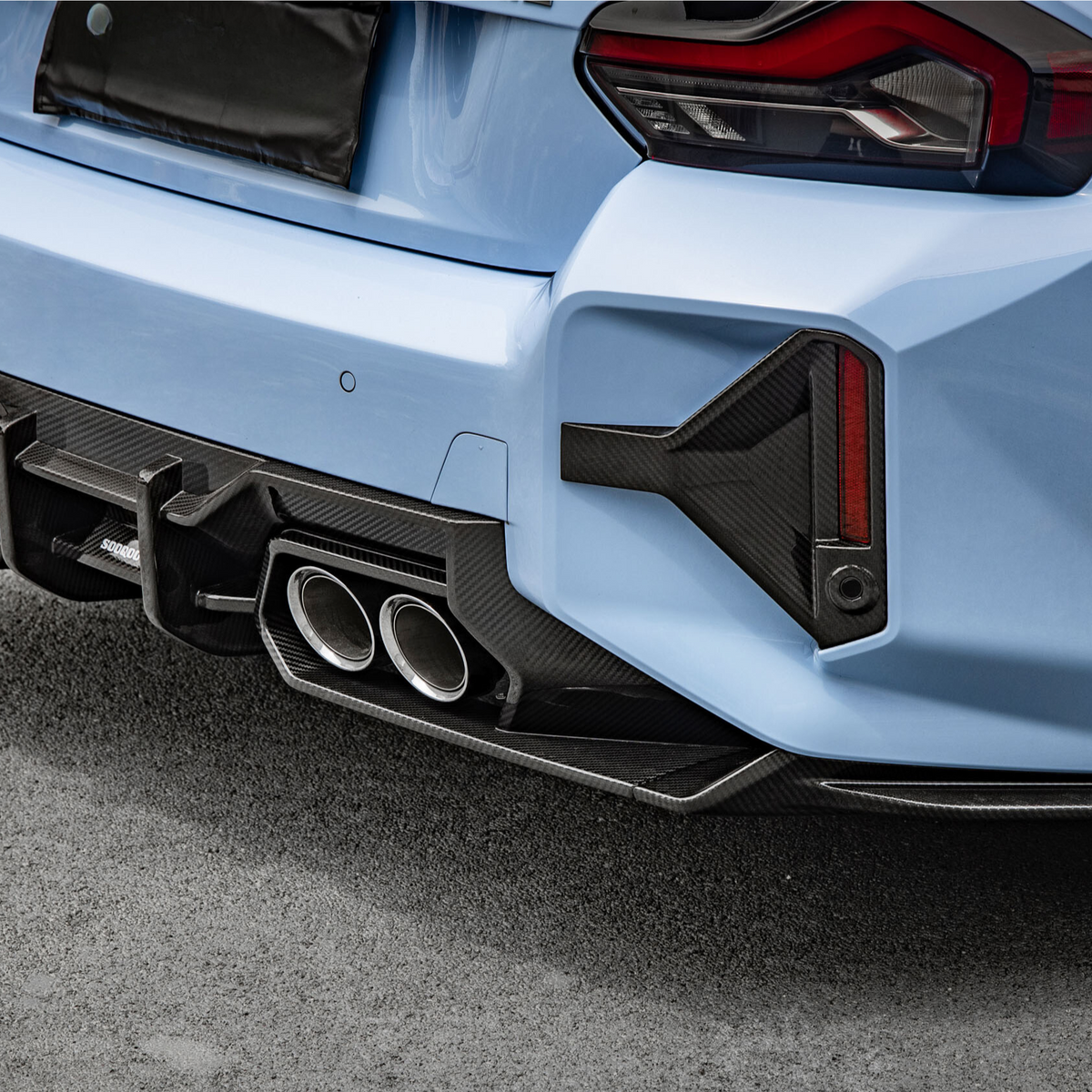 A light blue car with sporty dual exhaust pipes and a carbon fiber diffuser is parked on a paved surface. The rear taillights have an angular design.